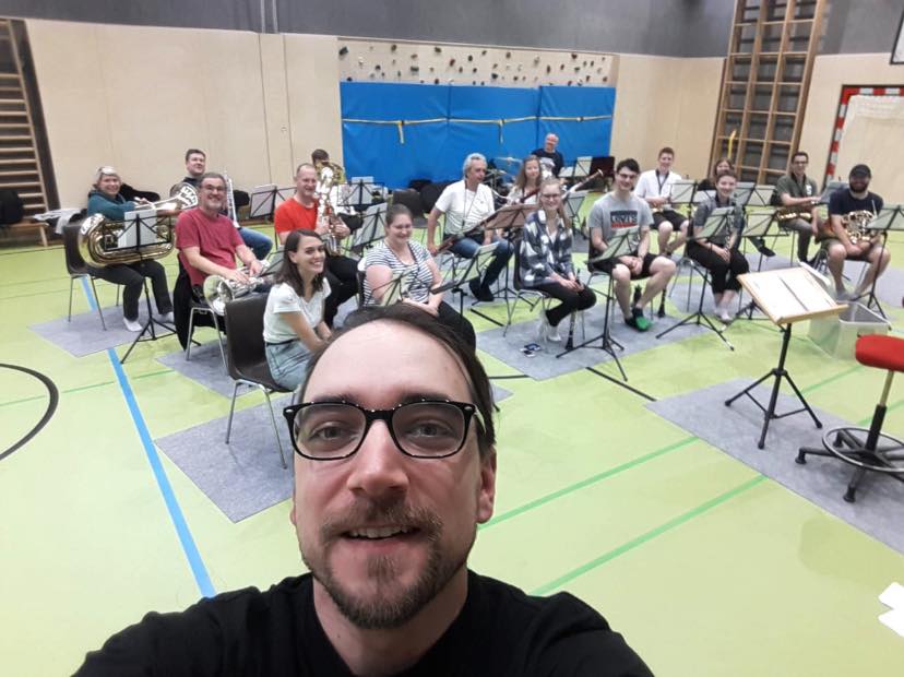 Ein Gruppenfoto aus der Probe im Turnsaal der Volksschule Aschach. Unser Kapellmeister Dizi steht im Fordergrund, die restlichen Musikerinnen und Musiker sitzen hinter ihm an ihren Notenständern. Wir lachen in die Kamera. 