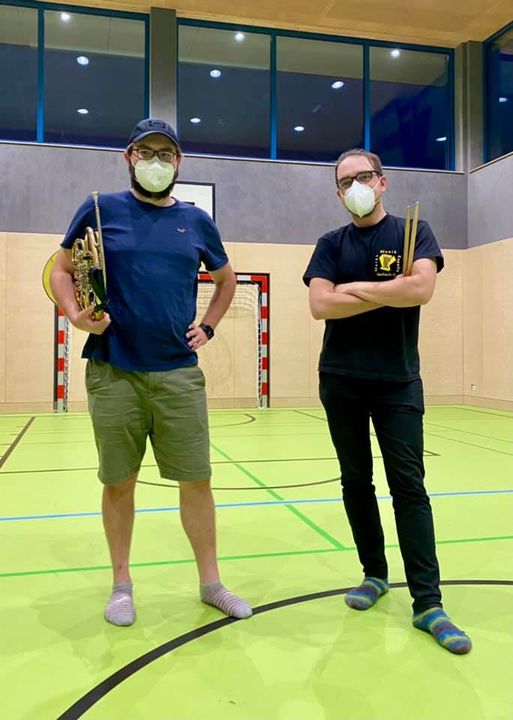 Das Bild zeigt unsere Musiker Augi und Dizi. Sie stehen im Turnsaal der VS Aschach und halten ihre Instrumente, Horn und Drumsticks, in den Händen.