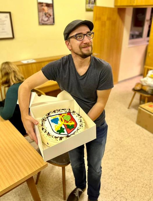 Das Bild zeigt unseren Kapellmeister Dizi, der zur Feier des Tages eine Torte mit dem Wappen von Aschach in der Hand hält. Das Foto ist im Probelokal aufgenommen worden.