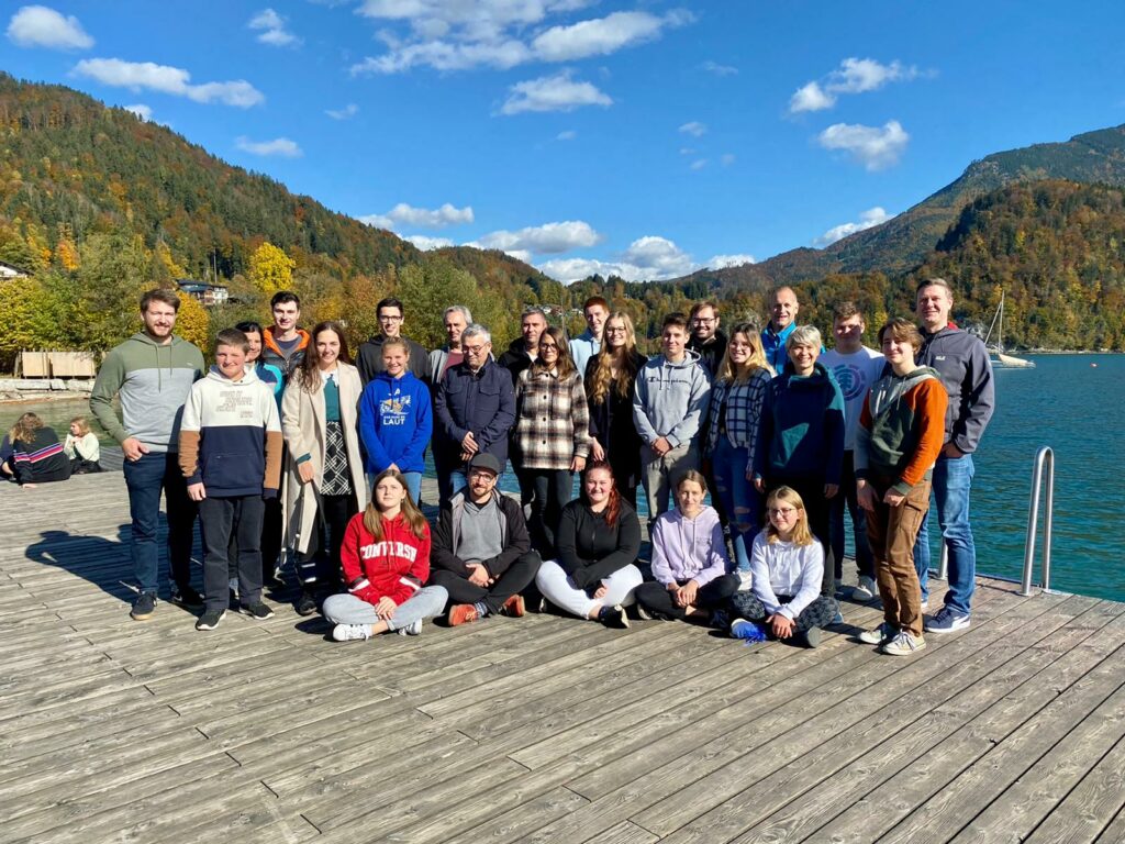 Ein Gruppenfoto der Musikerinnen und Musiker, die am Probewochenende dabei waren. Wir stehen am Steg in Sankt Gilgen am türkisblauen Wolfgangsee. Die Sonne strahlt uns ins Gesicht und wir lachen in die Kamera.